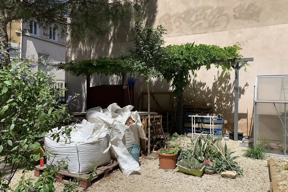 Jardin partagé en ville : bacs de culture, pergola couverte de vigne, sacs de terreau et plantes en pots au pied d'immeubles