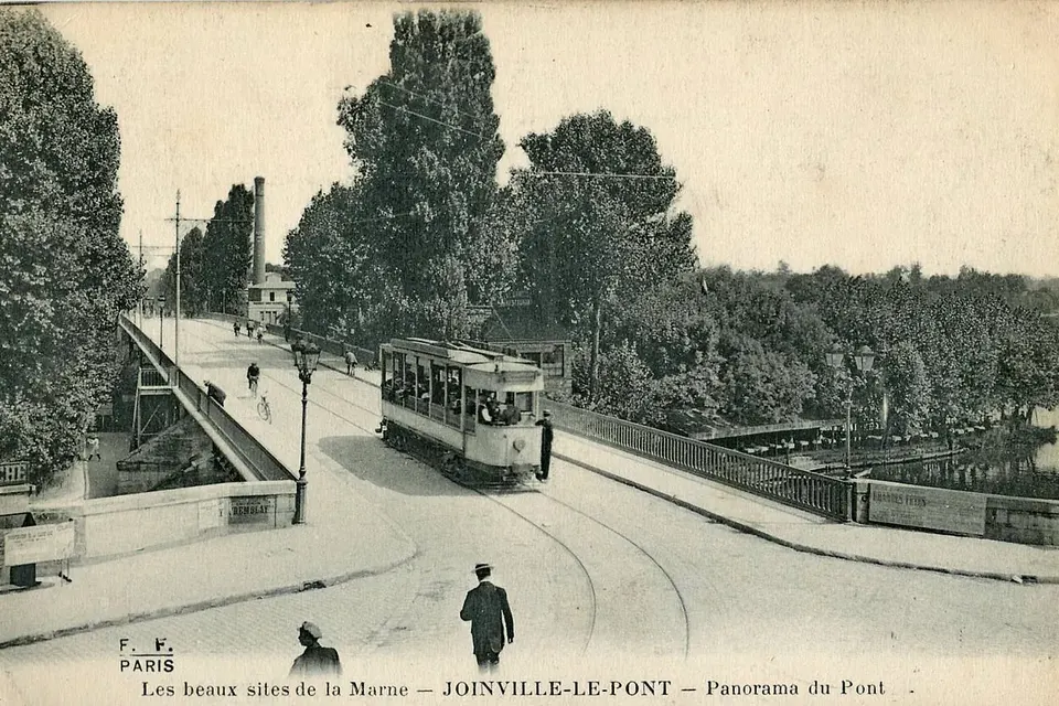 Carte postale ancienne : un tramway traverse le pont de Joinville-le-Pont, des passants sur le trottoir, des arbres le long de la Marne
