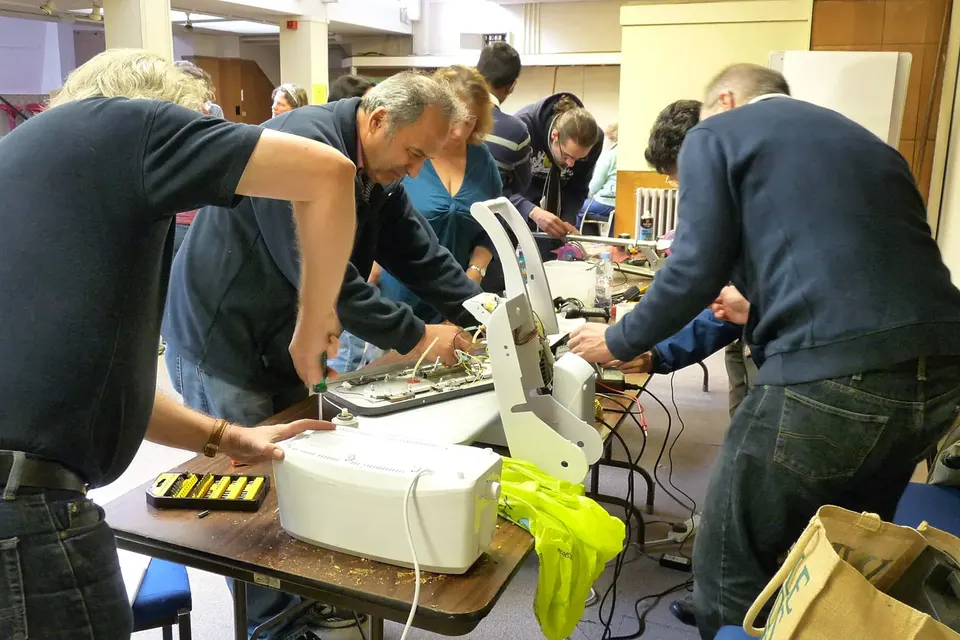 Des bénévoles penchés sur une table de travail réparent ensemble de petits appareils électriques, outils et boîtes à vis étalés