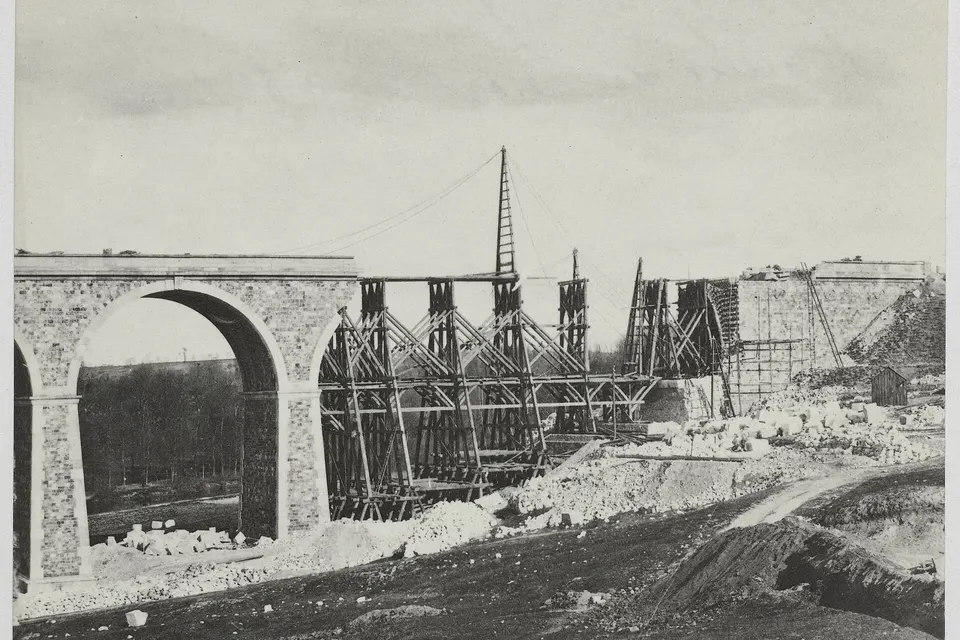 Photographie ancienne en noir et blanc : arches de pierre d'un viaduc ferroviaire en construction, échafaudages et grue en bois